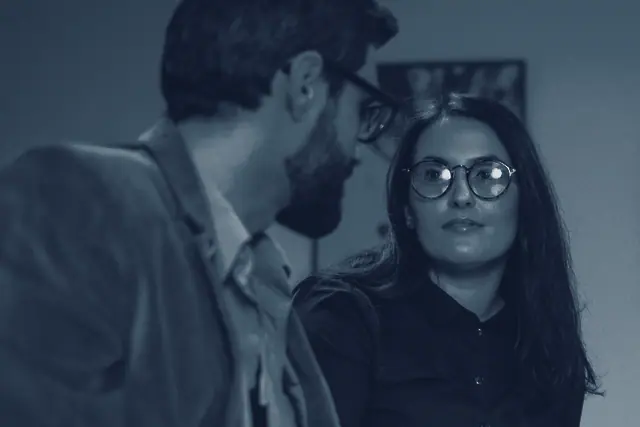 Un homme et une femme discutent assis à une table ; elle l'écoute en le regardant.