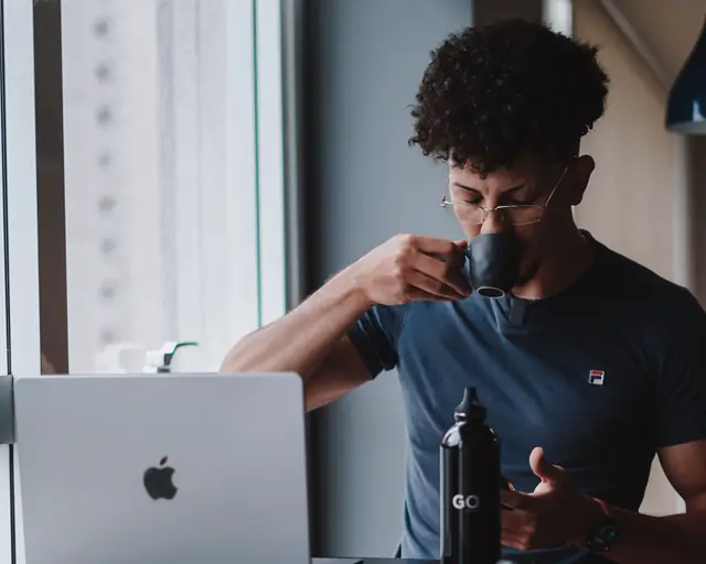 Un home pren cafè davant del portàtil, al costat d'una finestra.